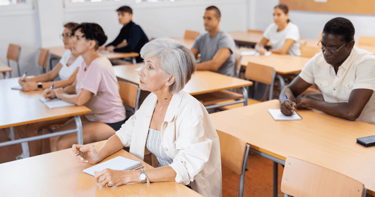 Adult students of different ages and backgrounds in a language class, writing in notebooks and facing the front of the room.