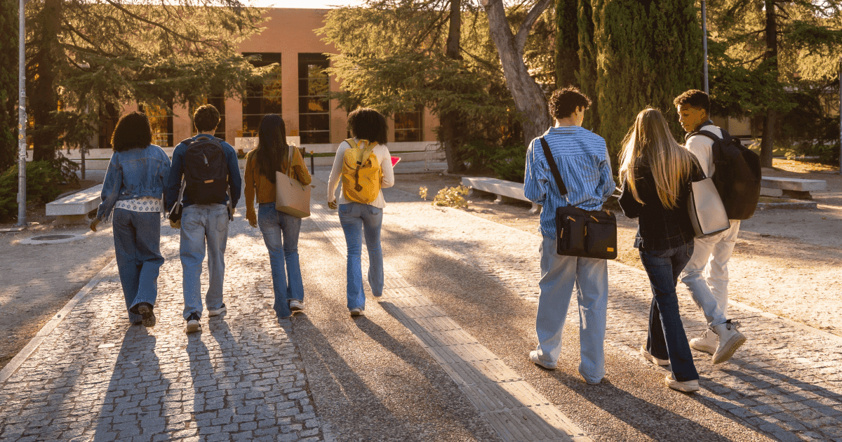 Six students walking together on a cobblestone campus path towards a building, seen from behind, with backpacks and bags, in warm golden-hour light.
